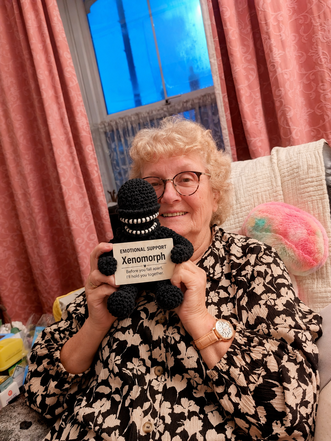 An older woman with glasses and blonde hair sits in a chair holding a black crocheted monster toy with a sign that reads 'Xenomorph'. The background shows pink curtains and a window.